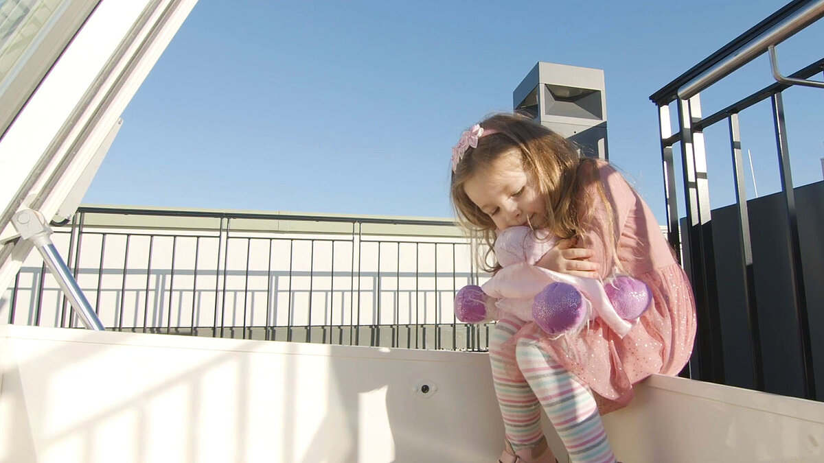 Kleines Mädchen mit Stofftier sitzt am geöffneten LAMILUX Flachdach Ausstieg und genießt die Sonne.