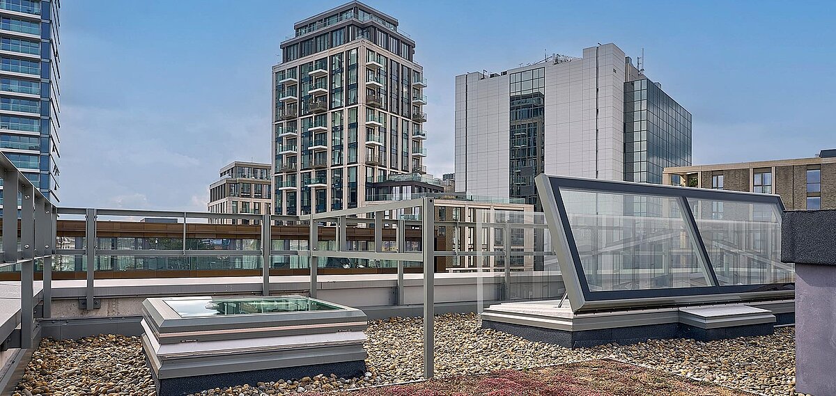 Dachfläche der Mulberry Academy London mit LAMILUX Tageslichtsystemen, Kiesbelag und Blick auf umliegende moderne Gebäude.