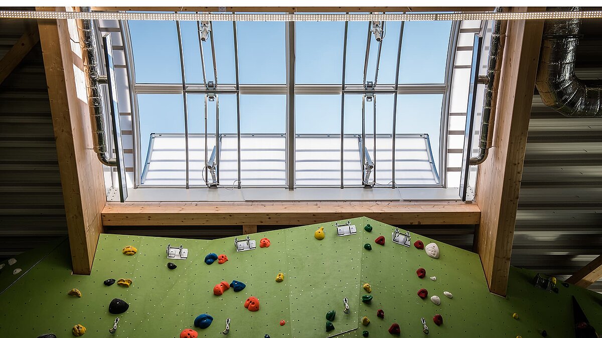 Blick auf Lichtbänder im Kletterzentrum OWL, darunter eine grüne Kletterwand mit bunten Klettergriffen.