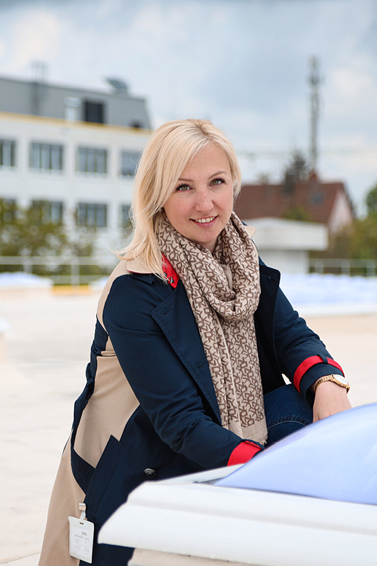 Frau mit hellem Schal und dunkler Jacke im Außenbereich, lächelnd an eine Lichtkuppel auf dem Dach angelehnt, modernes Gebäude im Hintergrund