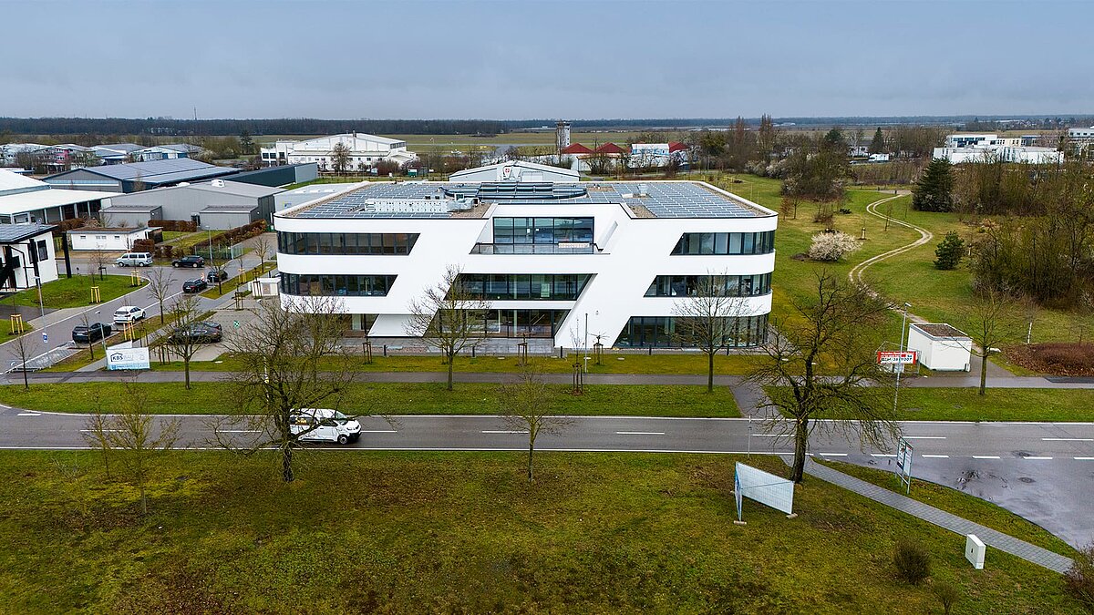 Modernes Bürogebäude mit weißer Fassade und Solaranlage auf dem Dach, umgeben von Grünflächen und Straßen, bei bewölktem Himmel.