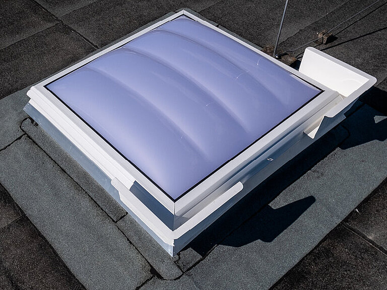 Quadratische Lichtkuppel auf Flachdach mit gewölbter Verglasung und weißem Aufsatzrahmen zur natürlichen Tageslichtversorgung