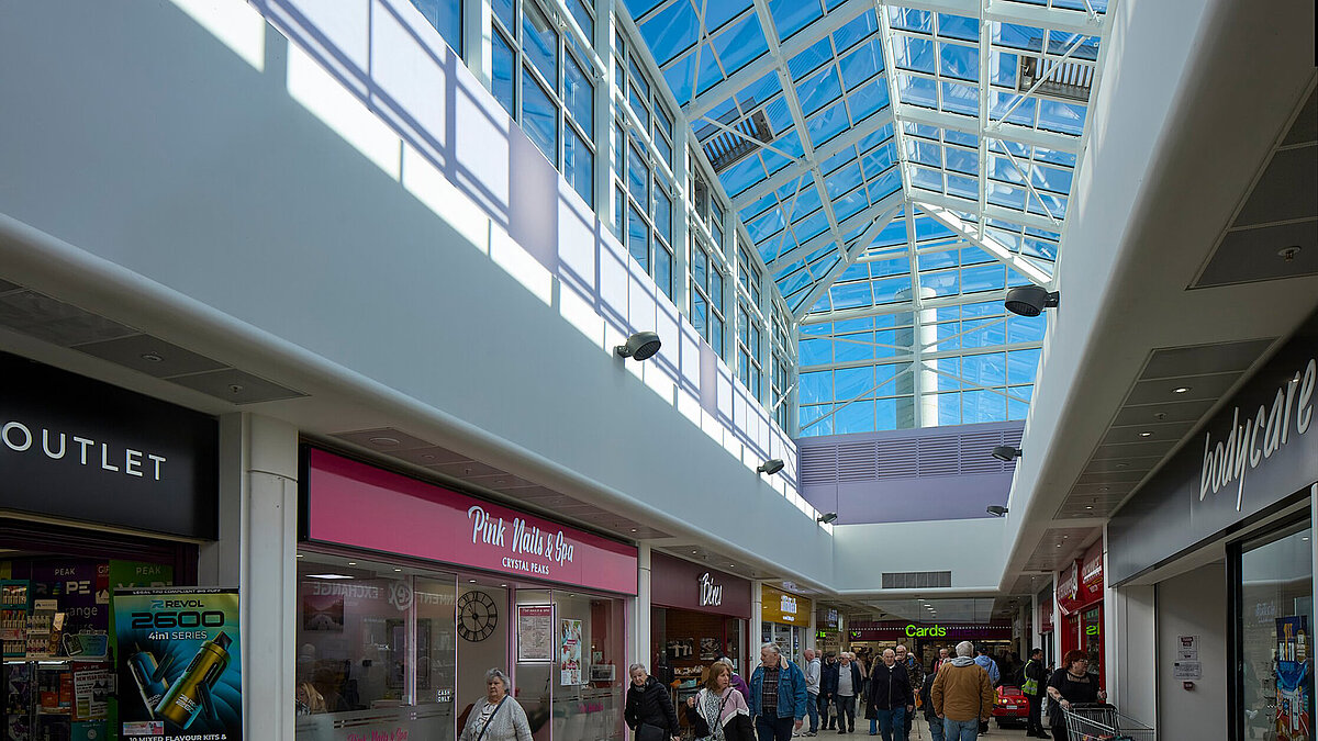LAMILUX Glasdach mit blauer Metallstruktur über einem Einkaufszentrum mit Geschäften und Personen, Tageslicht
