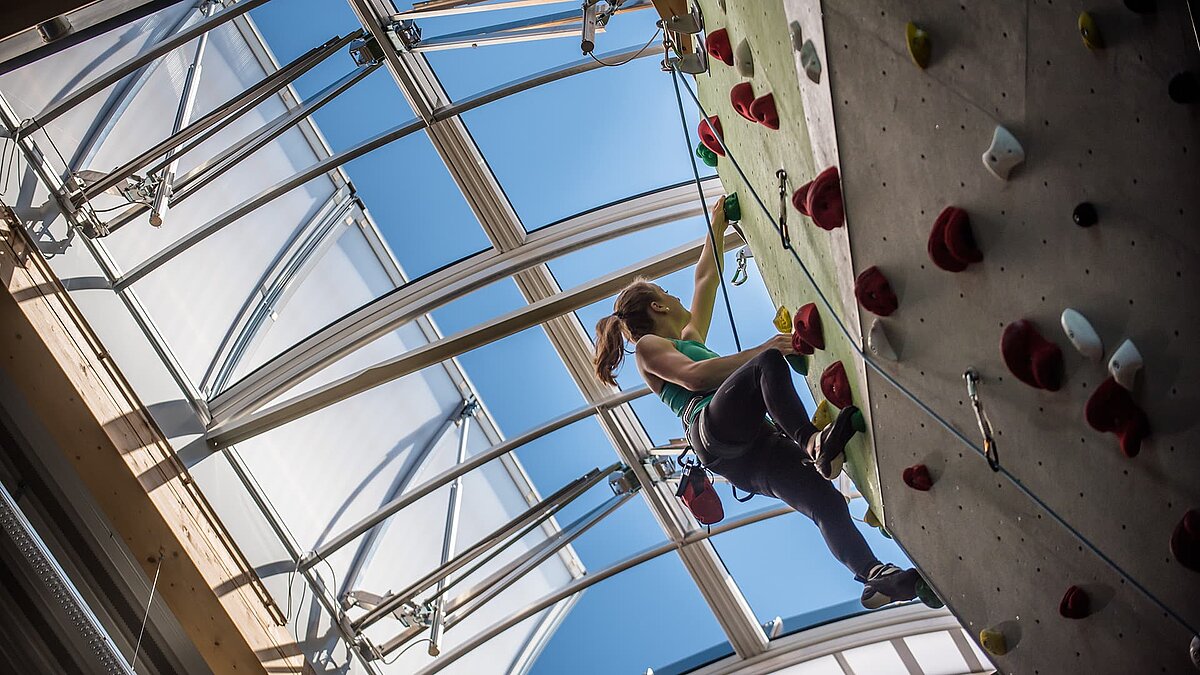 Kletterin an einer Indoor-Kletterwand im Kletterzentrum OWL unter einer großen Lichtkuppel, die natürliches Tageslicht hereinlässt.