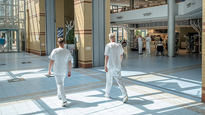 Zwei Klinikmitarbeiter gehen durch lichtdurchflutetes Krankenhausatrium mit Glasdach.