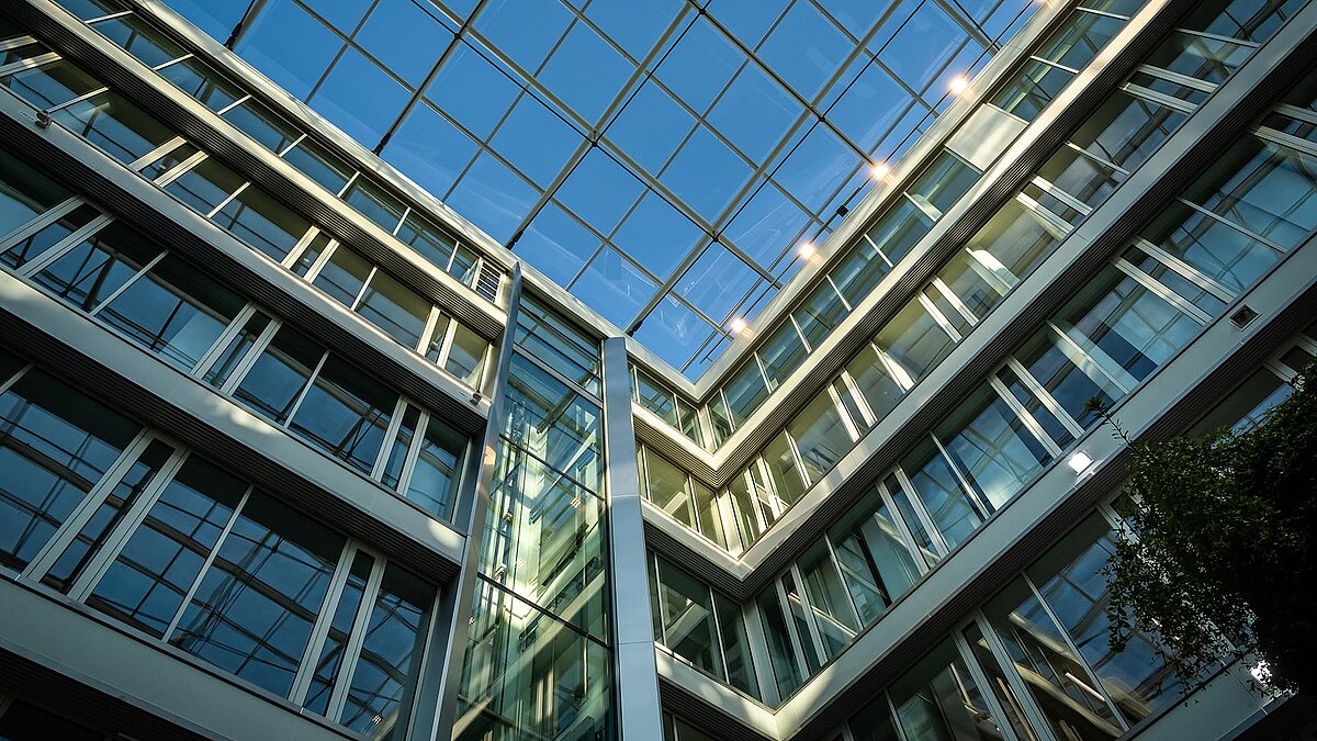 Blick nach oben auf das moderne Glasdach und die Fassaden des Bürogebäudes Newton der TÜV Süd Gruppe in München, mit reflektierenden Fenstern und blauem Himmel.