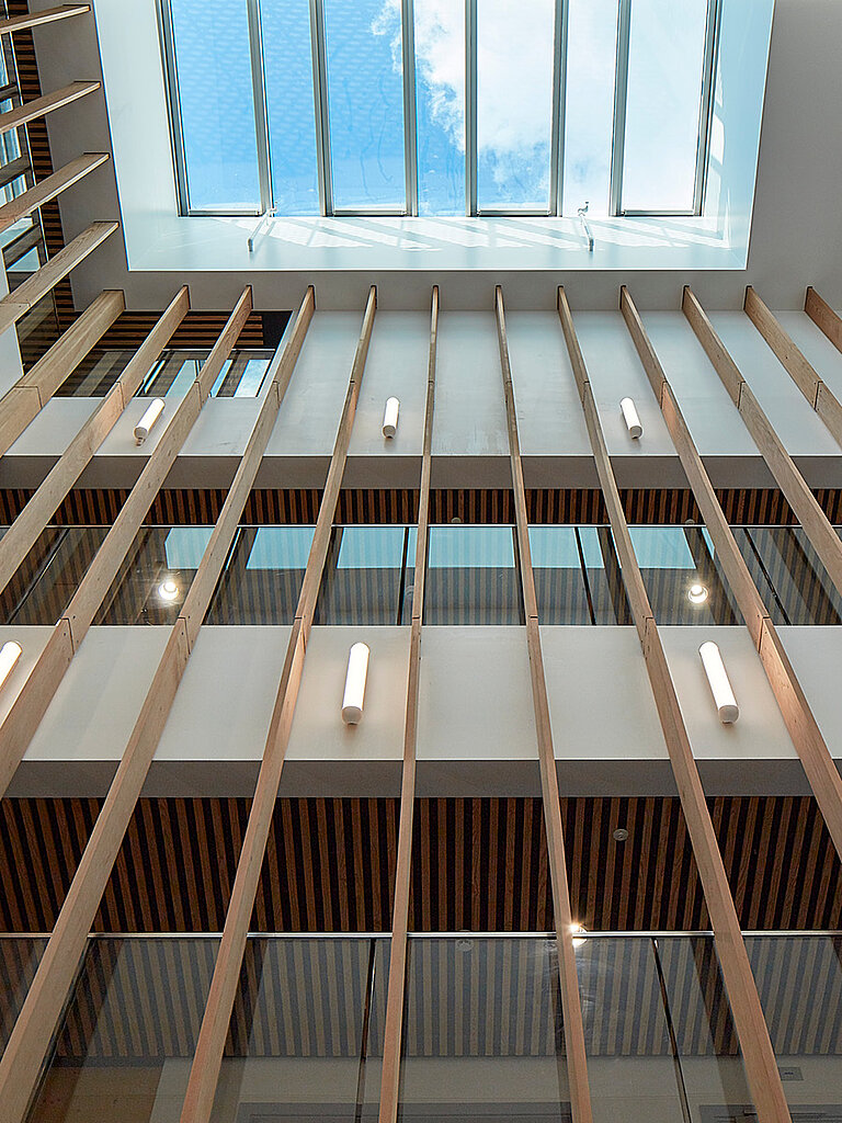 Blick nach oben in einen mehrgeschossigen Innenraum mit vertikalen Holzlamellen, Galerieebenen und zentralem Dachoberlicht mit Tageslichteinfall