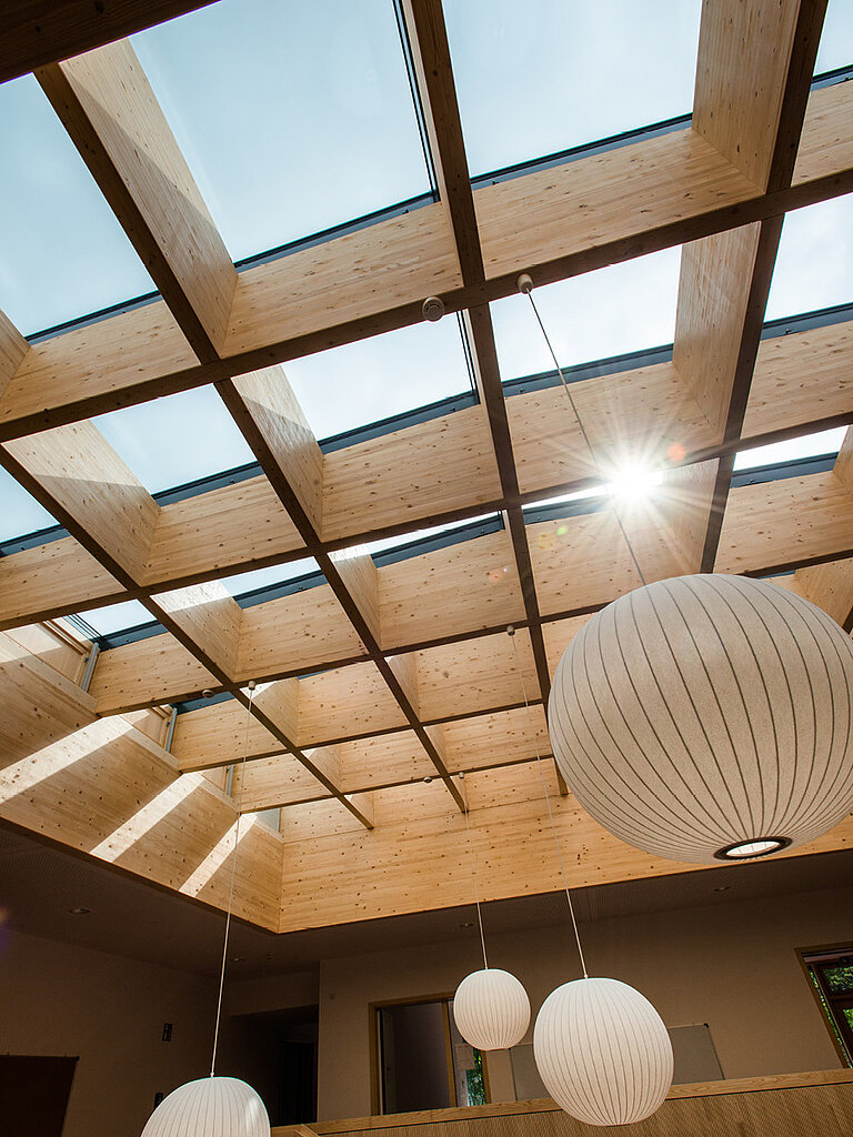 Innenansicht eines modernen Raums mit großflächiger Dachverglasung, Holztragstruktur und hängenden Leuchtelementen mit natürlichem Tageslichteinfall