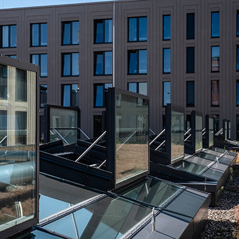 Mehrere moderne Glasdach-Elemente mit integrierter Lüftungsflügel  auf einem Flachdach vor einer zeitgenössischen Bürogebäudefassade.