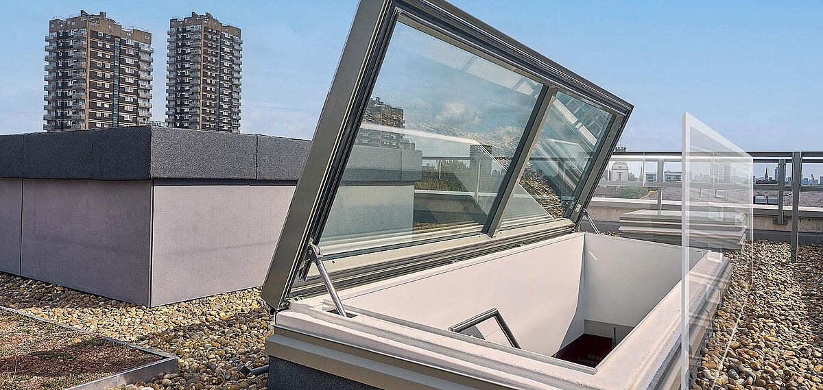Geöffneter LAMILUX Flachdach Ausstieg auf einem begrünten und mit Kies belegten Dach, im Hintergrund Hochhäuser und Skyline von London.