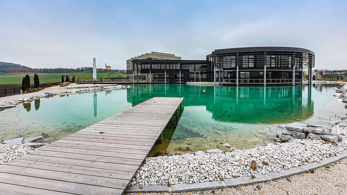 Hauptgebäude von Zaltech in Moosdorf, Österreich, mit einem großen Teich und Steg im Vordergrund.