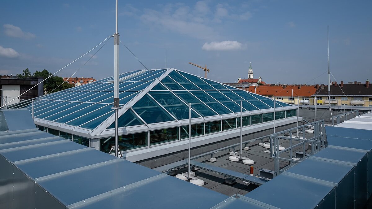 Glasdach des Dante-Gymnasiums mit stabiler Stahlkonstruktion und blauer Verglasung, umgeben von Gebäuden und blauer Himmel im Hintergrund.