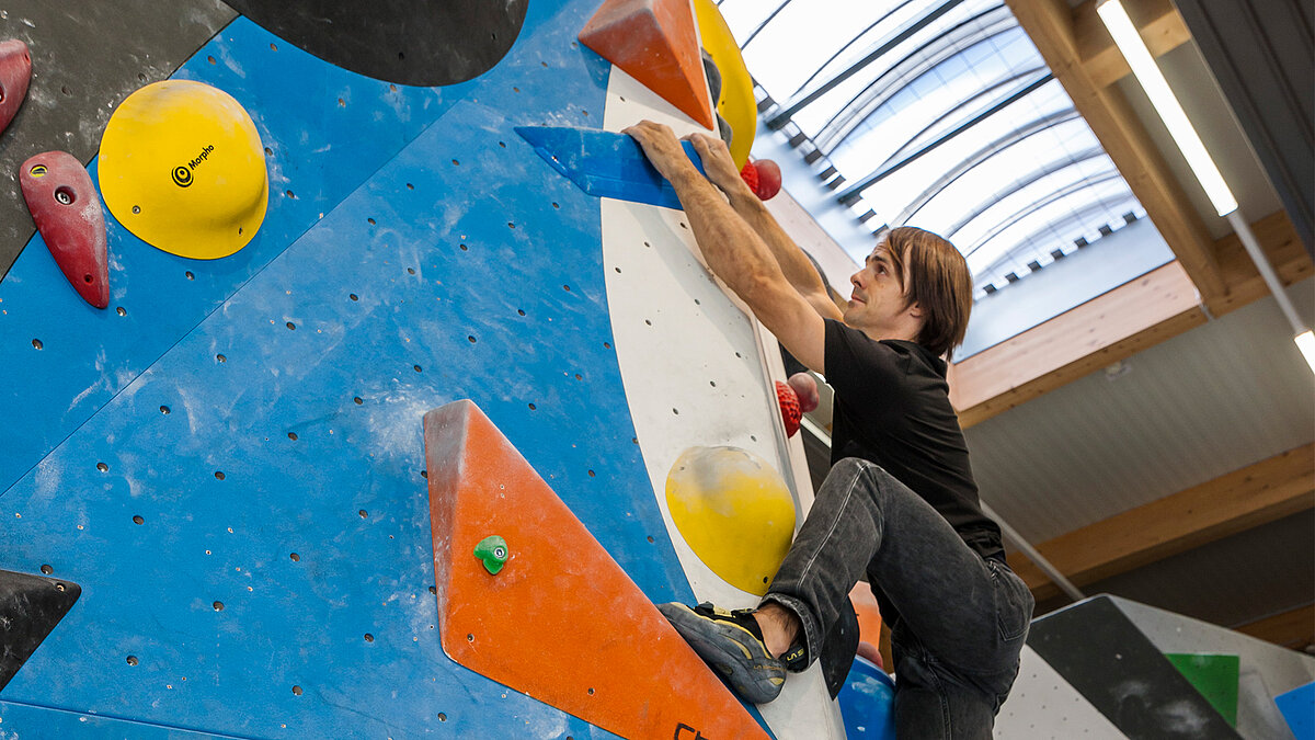 Mann in schwarzem Outfit klettert an einer Boulderwand in einer Kletterhalle