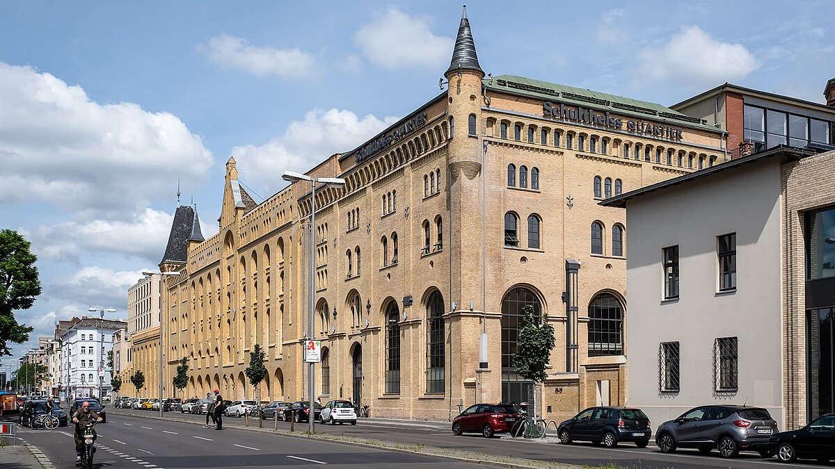 Außenansicht des Schultheiss Quartiers in Berlin-Moabit, historisches Backsteingebäude mit modernen Anbauten, Straßenszene mit Autos und Radfahrern.