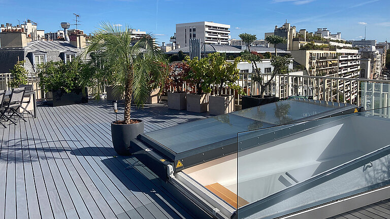 Moderne Dachterrasse in Paris mit Holzdielen, Palme, Kübelpflanzen, Glasgeländer und geöffnetem Flachdach Ausstieg