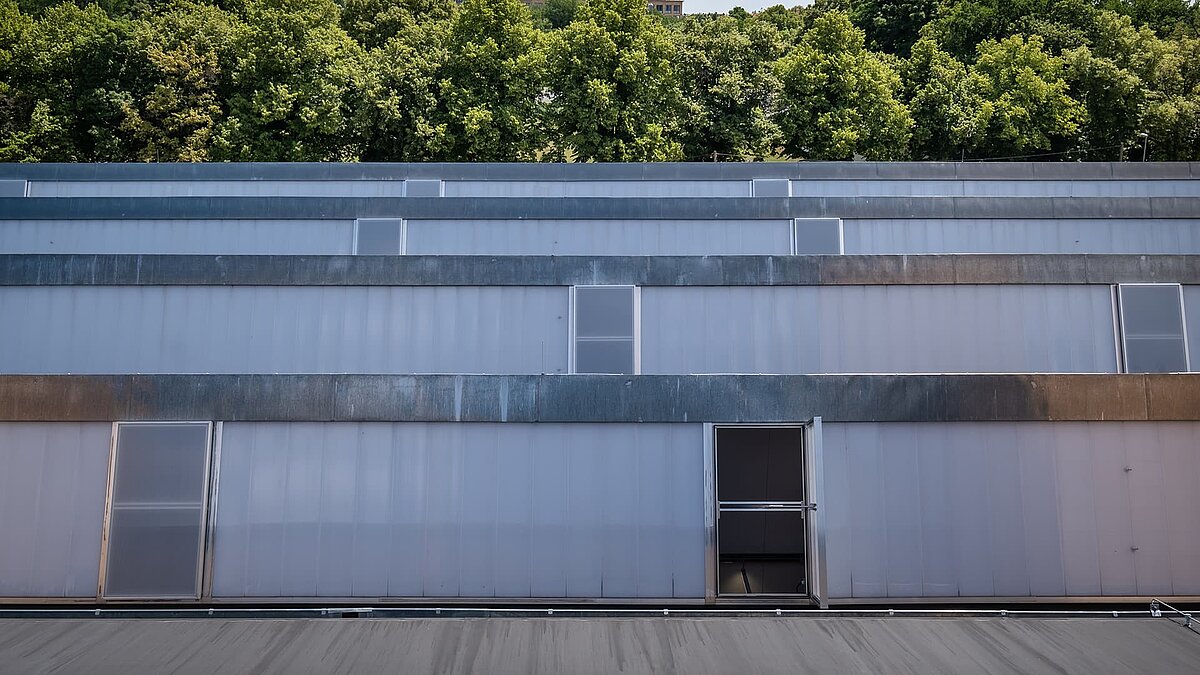 Außenansicht des alten Postbahnhofs Leipzig mit modernen Lichtbändern und teilweise geöffneten Fenstern, umgeben von grünen Bäumen.