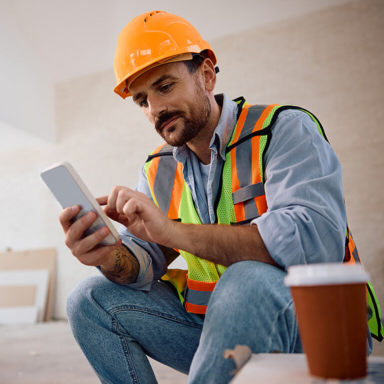 Bauarbeiter mit Schutzhelm und Warnweste nutzt Smartphone auf der Baustelle