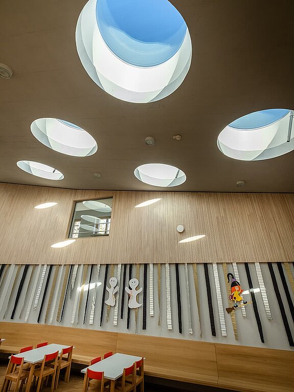 Innenraum des Kindergartens St. Severin mit runden Dachfenstern, Holzwänden, bunten Dekorationen und Kindertischen.