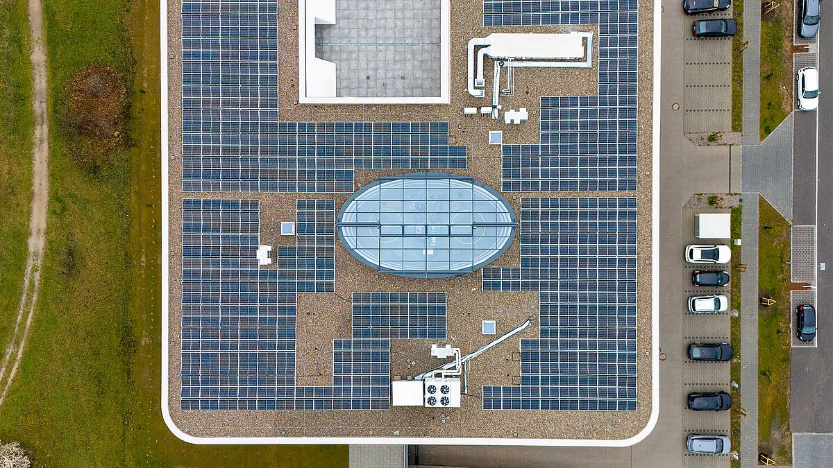 Drohnenaufnahme eines Flachdachs mit zahlreichen Solarpaneelen und einem ovalen Glasdach in der Mitte, daneben ein Parkplatz mit Autos.