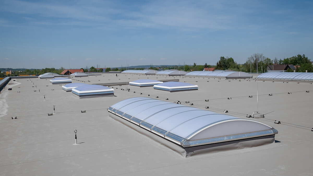 Dach der Produktionshalle des Bauunternehmens Unglehrt mit mehreren LAMILUX Lichtkuppeln F100 W und Lichtbändern B unter blauem Himmel.