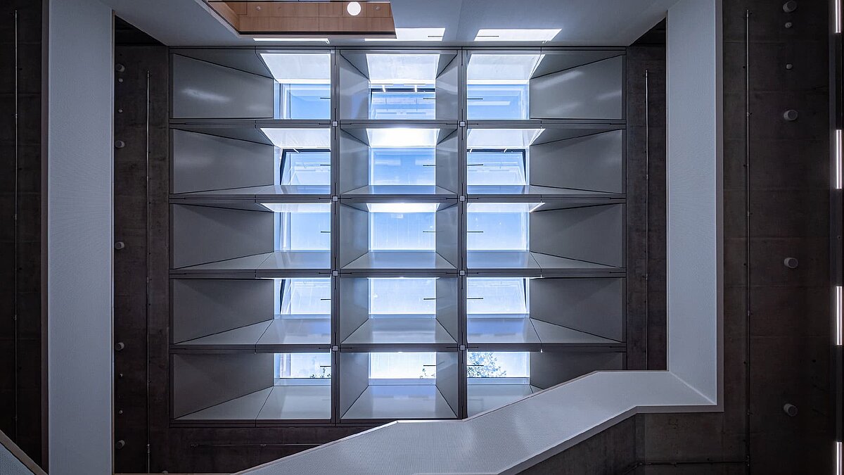 Blick nach oben auf ein quadratisches Glasdach mit umlaufenden Regalen in einem Innenraum