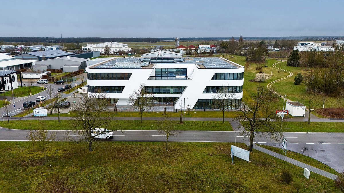 Modernes Bürogebäude mit weißer Fassade und Solaranlage auf dem Dach, umgeben von Grünflächen und Straßen, bei bewölktem Himmel.