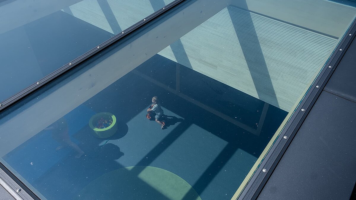 Blick durch das großflächige Glasdach der Kinderkrippe Wiggensbach auf spielende Kinder und Raum mit Tageslichteinfall.