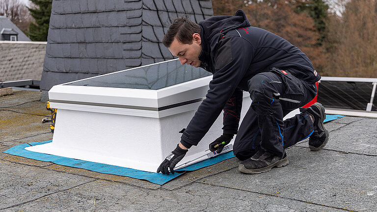 Handwerker in Arbeitskleidung montiert weißes Flachdach Fenster auf einem Flachdach mit blauer Abdichtungsfolie