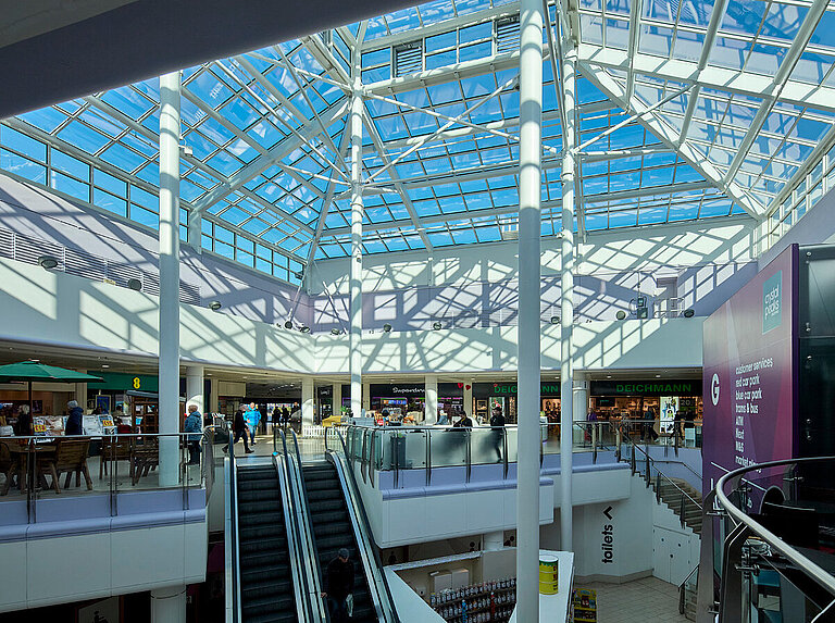Innenansicht eines Einkaufszentrums mit großem Glasdach, Tageslicht und Rolltreppen